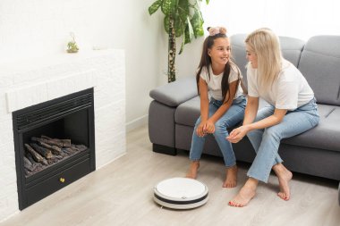 Cute child near mother looking at robotic vacuum cleaner in living room.