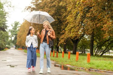 Cheerful mother and her little daughter having fun together in the autumn background under the umbrella. Happy family in the fall background.