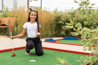 Ailesiyle mini golf oynayan tatlı bir anaokulu kızı. Yeni yürümeye başlayan çocuk dışarıda eğleniyor. Çocuklar ve yetişkinler için açık havada yaz sporu. Aile tatili ya da tatil yeri.