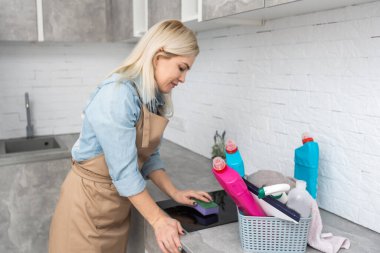 İnsanlar, ev işleri ve ev işleri konsepti. Evde mutfağı süngerle temizleyen bir şişe sprey ile sarışın orta yaşlı kadın.