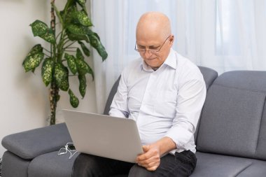 Beyaz gömlekli ve siyah pantolonlu yaşlı kel bir adam gözlük takıyor ve oturma odasında otururken dizüstü bilgisayarla çalışıyor.