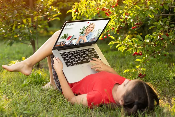 Beautiful young lady is chatting with friends outdoors as it is very useful to be in nature. Laptop is the best friend for a child nowadays. Green and vast meadow.