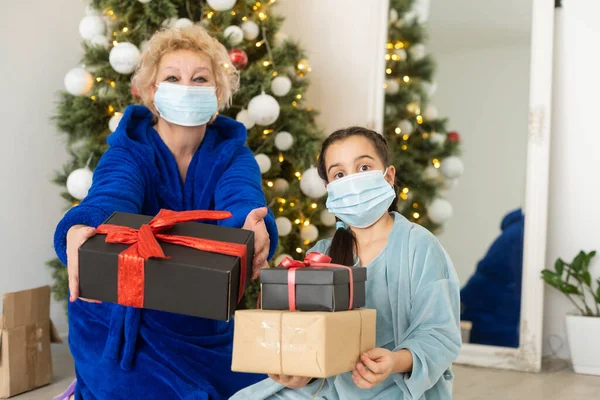 Grandmother and granddaughter in the living room with Christmas decor hugging in medical masks on their faces. A family holiday during the outbreak of coronavirus and disease. New Year.