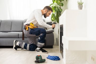 Professional technician with screwdriver installing electric fireplace in room.
