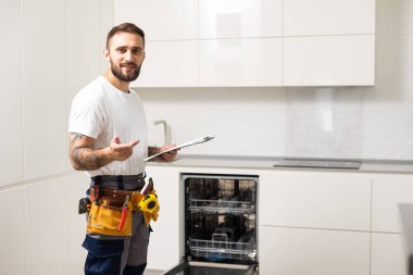 Ev mutfağındaki bulaşık makinesinin tamiri sırasında işçi kıyafeti giymiş genç modern bir tamirci..