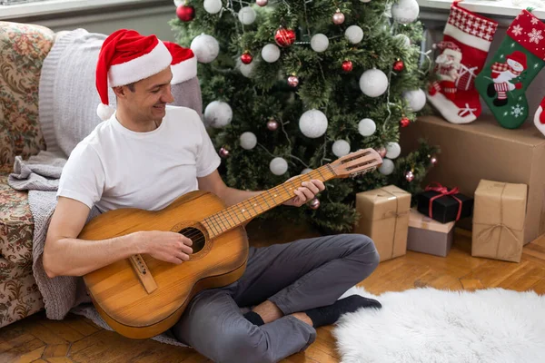 幸せな若い男がギターを弾いている 男は喜んで見ていると屈託のない クリスマスや新年を祝うだけでお祝いの帽子の男性 背景にガーランドとクリスマスツリー — ストック写真