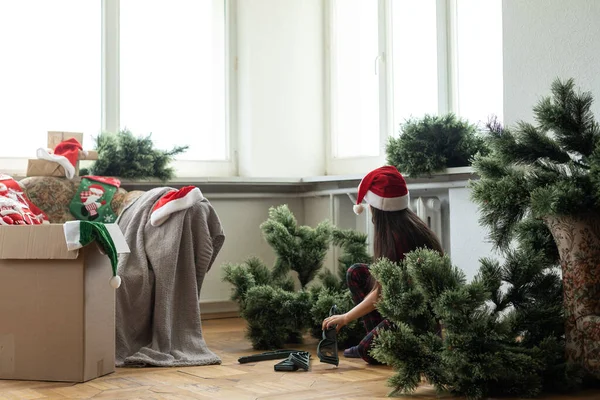 かわいい陽気な女の子の写真人工的なクリスマスツリーは屋内でサンタの帽子を着用します — ストック写真