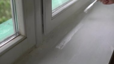 Bir kadın pencerenin yamaçlarını beyaz boyayla boyuyor, pencere açıklarını boya fırçasıyla boyuyor, pencere çerçevelerini beyaz boya ile boyuyor.