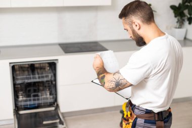 Ev mutfağındaki bulaşık makinesinin tamiri sırasında işçi kıyafeti giymiş genç modern bir tamirci..