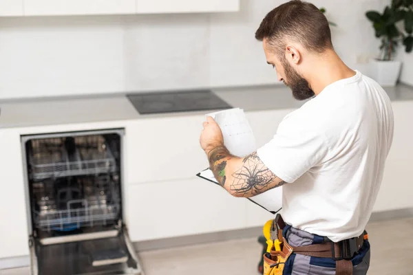 Young modern serviceman in worker suit during the repairing of the dishwasher on the domestic kitchen.