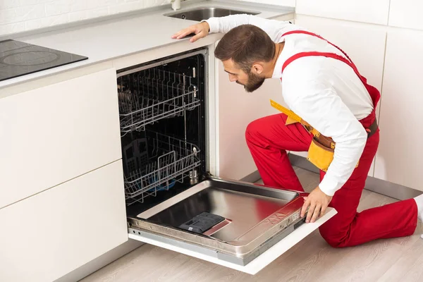 Young modern serviceman in worker suit during the repairing of the dishwasher on the domestic kitchen.