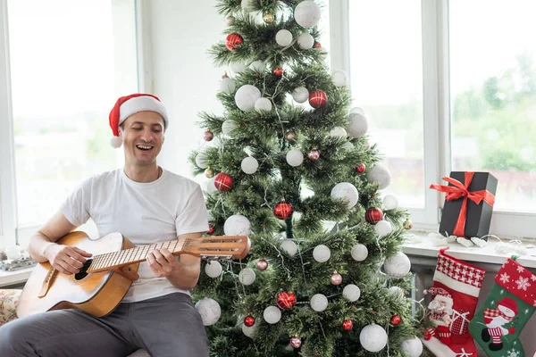 幸せな若い男がギターを弾いている 男は喜んで見ていると屈託のない クリスマスや新年を祝うだけでお祝いの帽子の男性 背景にガーランドとクリスマスツリー — ストック写真