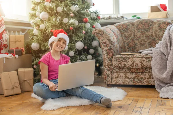 幸せな女の子は自宅でクリスマスツリーの下に贈り物を開き ビデオリンクを介してラップトップを介して祖父母と通信します クリスマスと新年のための休日の概念 — ストック写真