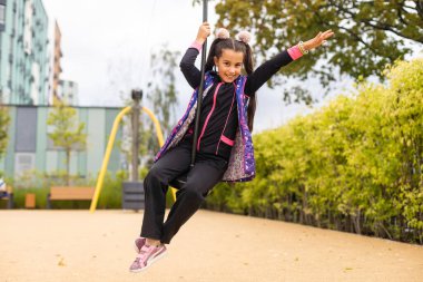 Gülümseyen küçük kız oyun parkında ipte sallanıyor.