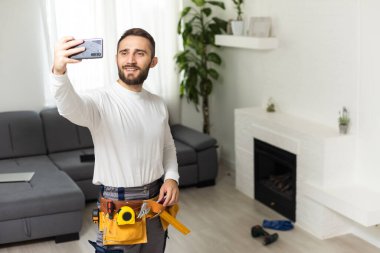 Yakışıklı, neşeli tamircinin portresi. Akıllı telefondan selfie çekiyor. Gömlek ve tulum giyiyor..