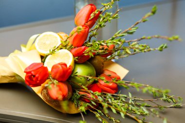 Mevsimlik meyve tabağı. Beyaz tabakta limon ve elma. Kırmızı lale çiçekleri ve yeşil yapraklı dallar.