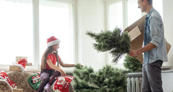 娘を持つ幸せな親は自宅のリビングルームにクリスマスツリーを設置し — ストック写真