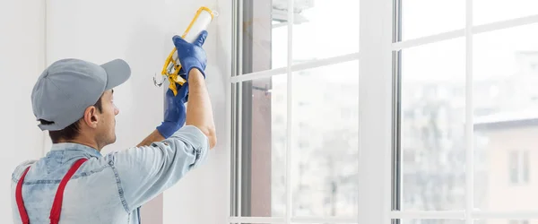 Construction worker sealing window with caulk indoors.