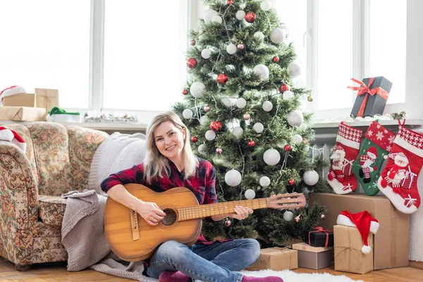 young woman holds a guitar in hand and sits in christmas studio.
