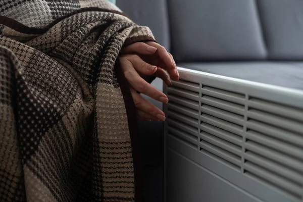 Girl warms up the frozen hands above hot radiator, close up. Woman wearing woolen sweater warming up while sitting near a heating radiator. Woman warming hands near electric heater at home