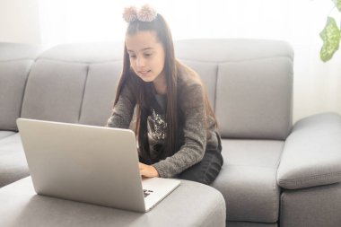 Kafkasyalı genç kız, elinde bilgisayarla evdeki gri koltukta oturuyor.