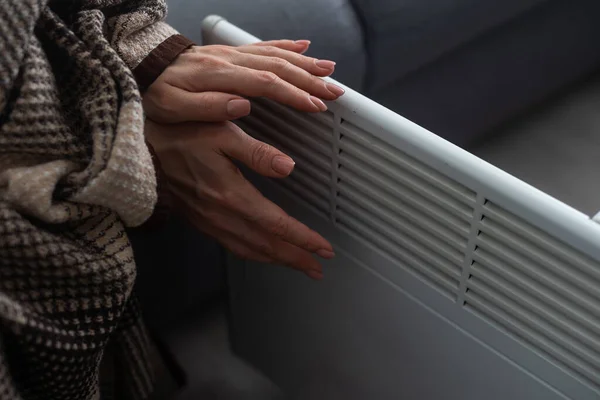 Girl warms up the frozen hands above hot radiator, close up. Woman wearing woolen sweater warming up while sitting near a heating radiator. Woman warming hands near electric heater at home