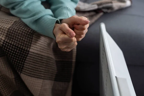 Girl warms up the frozen hands above hot radiator, close up. Woman wearing woolen sweater warming up while sitting near a heating radiator. Woman warming hands near electric heater at home