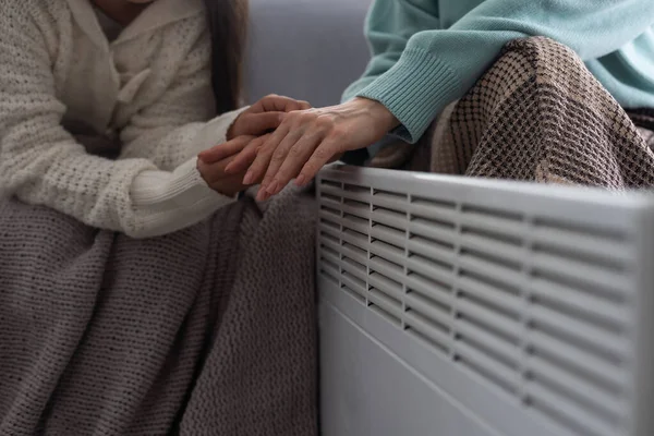 At the electric heater, people are warming themselves, covered with a warm blanket at home. hands of mother and daughter near the heater. Cold season and gas crisis in Europe.