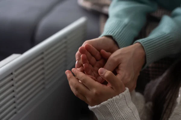 Mother and child warming hands near electric heater at home, closeup
