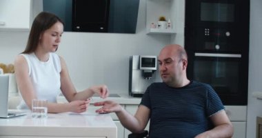 Kadın tekerlekli sandalyedeki adama bir bardak suyla ilaç veriyor. Engellilere yardım