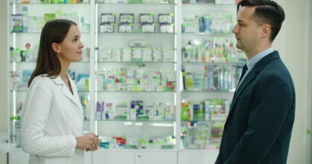 Konsultacje. Kaukaska młoda farmaceutka doradzająca lekom, tabletkom, lekom, lekom przeciwbólowym, witaminom klientowi płci męskiej, kupcowi w aptece — Wideo stockowe