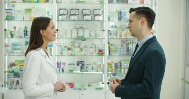 Konsultation. Kaukasische junge Apothekerin, die einem männlichen Kunden, einem Käufer in der Drogerie Medikamente, Pillen, Medikamente, Schmerzmittel, Vitamine berät — Stockvideo