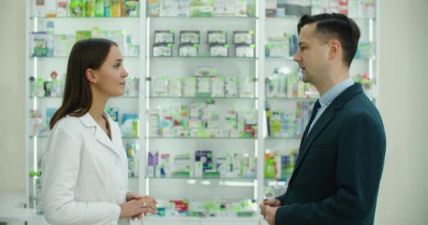 Konzultace. Kavkazská mladá farmaceutka, která radí lékům, práškům, lékům, lékům proti bolesti, vitamínům klientovi mužského zákazníka, kupujícímu v lékárně — Stock video