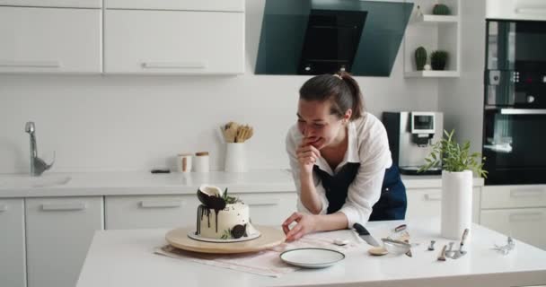 Banketbakker in de keuken maakt taart. Thuis aan het koken. Heerlijke en mooie zelfgemaakte taarten, banketbakkersrecepten. Een jonge vrouw in een schort in de keuken sneed de taart — Stockvideo