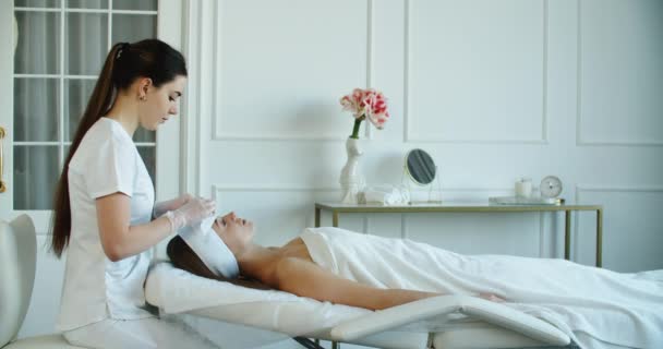 Cilt sorunlarını tedavi. Kadın kozmetik uzmanı müşterilerin yüzüne bakıp yüz masajı yapıyor. Mutlu, rahat, genç bir kadın spa salonunda profesyonel yüz bakımı görüyor. — Stok video