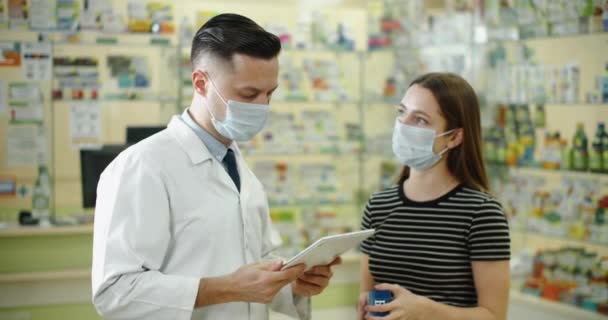 Orvosi maszkot viselő férfi gyógyszerész, aki orvosi maszkban szolgál fel egy ügyfelet egy gyógyszertárban, miközben megmutatja neki a digitális tablettát. Tanácsadás és segítség a világjárvány idején. — Stock videók