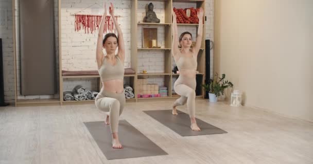 Zwei junge Frauen machen Yoga auf Matten und machen die Übungen synchron. Sie setzen sich und heben die Hände. — Stockvideo