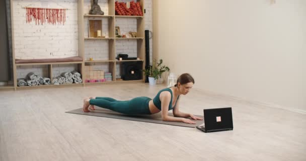 Exercício em um estúdio de Yoga. Menina desportiva fazendo prancha de ioga enquanto assiste tutorial on-line no laptop, espaço livre — Vídeo de Stock