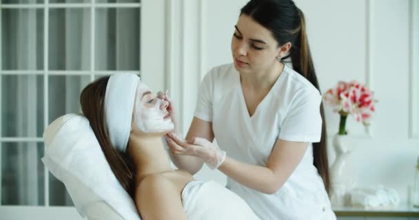Женщина-косметолог делает для клиента процедуры по уходу за лицом в клинике эстетической медицины. Косметолог или дерматолог делает пилинг или лифтинговый массаж женщине в спа или салоне. — стоковое видео
