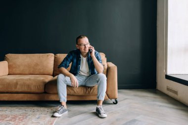 Gözlüklü bir adam gündelik giysiler içinde koltukta oturmuş gülümsüyor ve telefonda konuşuyor.