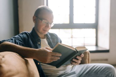Gözlüklü ve günlük kıyafetli bir adam pencerenin arkasındaki koltukta kitap okuyor.