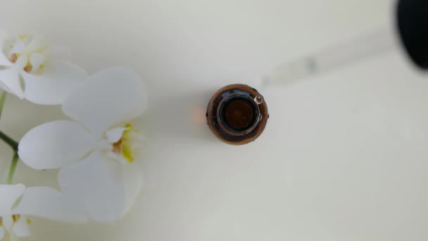 Glaskosmetikflasche mit Pipette liegt mit einer weißen Orchidee auf der Wasseroberfläche. Draufsicht auf Tropfen fällt ins Wasser und divergierende Wasserkreise auf weißem Hintergrund. Verpackungsmuster für Design, Werbung. 4k