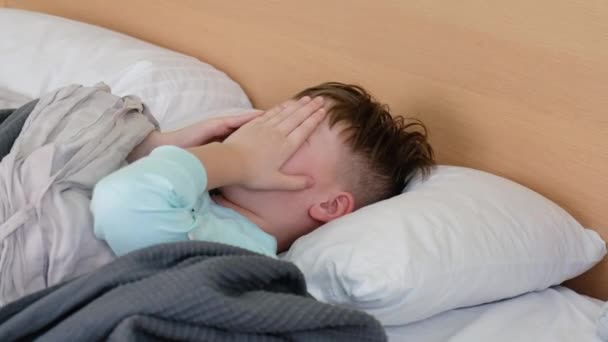 Ein trauriger Junge liegt mit Fieber und Kopfschmerzen auf dem Bett und reibt sich vor Schmerzen den Kopf. Das Gesicht ist vor Schmerz verdreht — Stockvideo