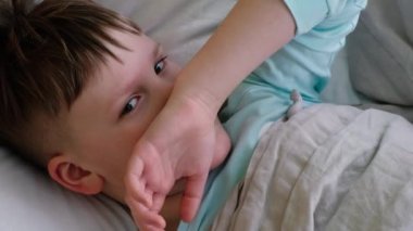 Üzgün bir çocuk ateşi ve baş ağrısı ile yatakta yatar ve başını acıdan ovuşturur. Yüzü acıdan burkulmuş.