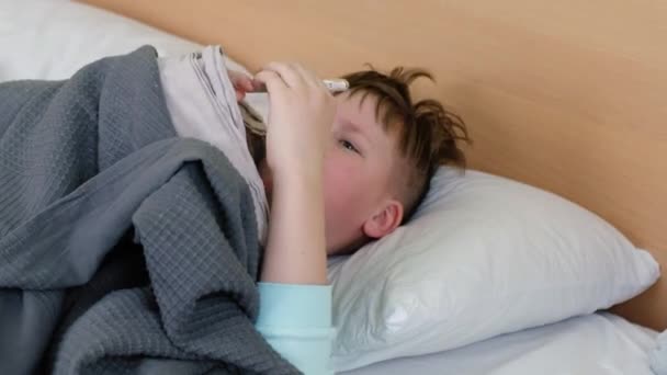 Грустный мальчик лежит на кровати и измеряет температуру ртутным термометром, и у него болит голова. Лицо искажено болью — стоковое видео