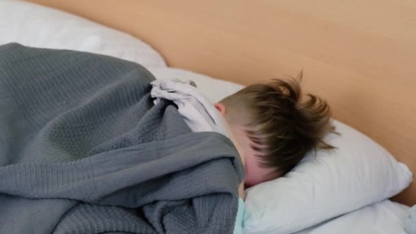 Seorang anak laki-laki yang sedih berbaring di tempat tidur dengan demam dan sakit kepala dan menggosok kepalanya kesakitan. Wajahnya terpelintir dengan rasa sakit — Stok Video