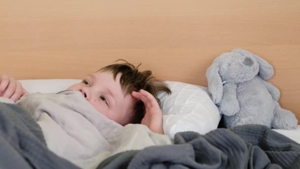 A sad boy lies on the bed with a fever and headache and rubs his head in pain. The face is twisted with pain — Stock Video