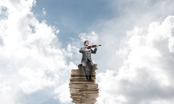 Guapo violinista en el cielo azul del día en un montón de libros toca su melodía —  Fotos de Stock