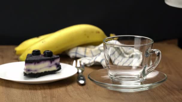 将热茶倒入透明的玻璃杯中 配上一块美味的自制蓝莓芝士蛋糕 并配上保存好的蓝莓 成熟的香蕉 木制桌子上的金属叉子 — 图库视频影像