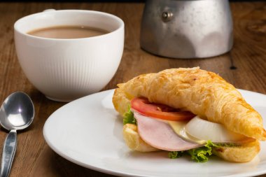 Kahvaltıda lezzetli jambonlu peynirli kruvasan ve sıcak sütlü kahve manzarası. Yeşil sebzeli jambonlu kruvasan ve beyaz seramik tabakta domates..